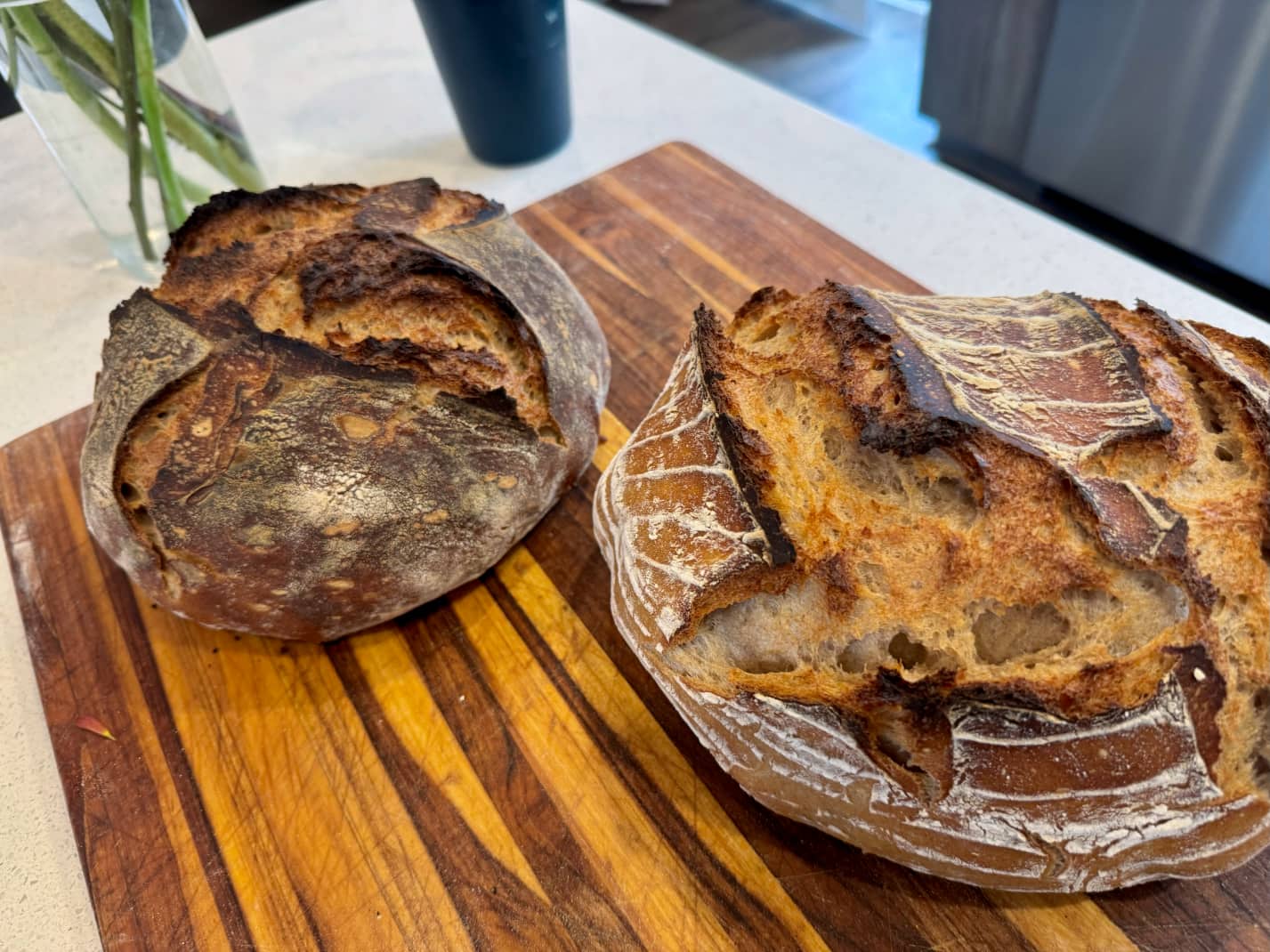 Two loaves of sourdough bread, one round and one batard-like, both with dark brown crusts and sliced tops, on a teak cutting board